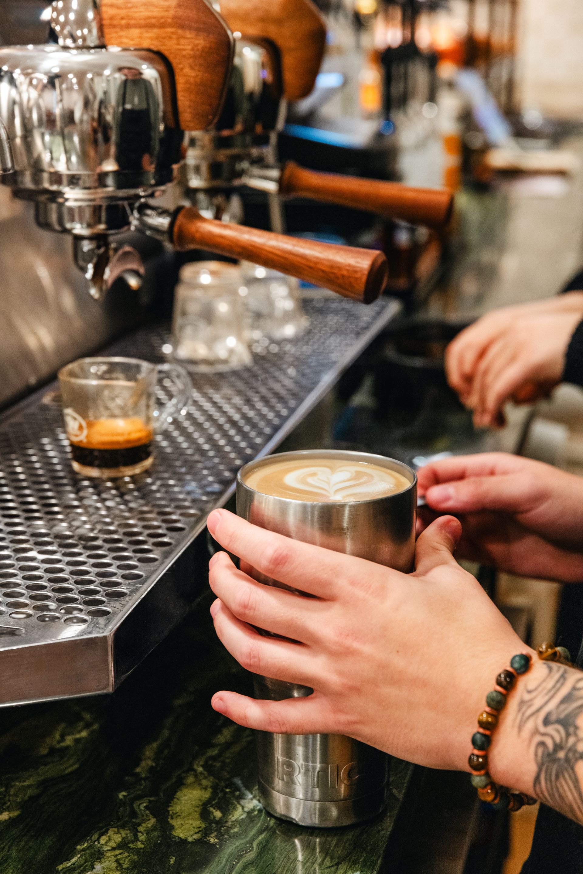 Hands holding a metal cup at a café espresso machine, with coffee being poured nearby.