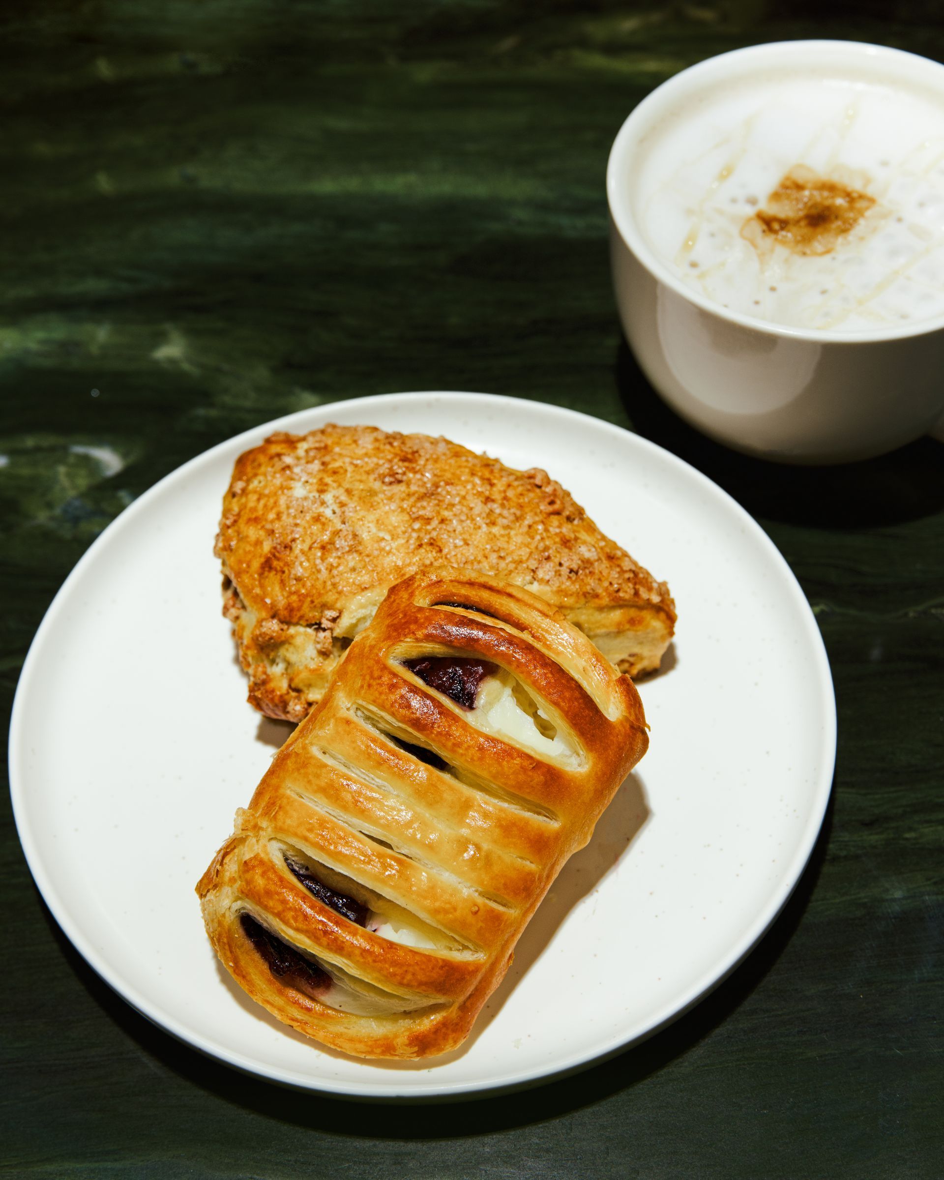 White plate with a glazed pastry and a sliced chocolate pastry, beside a cup of coffee on a dark table