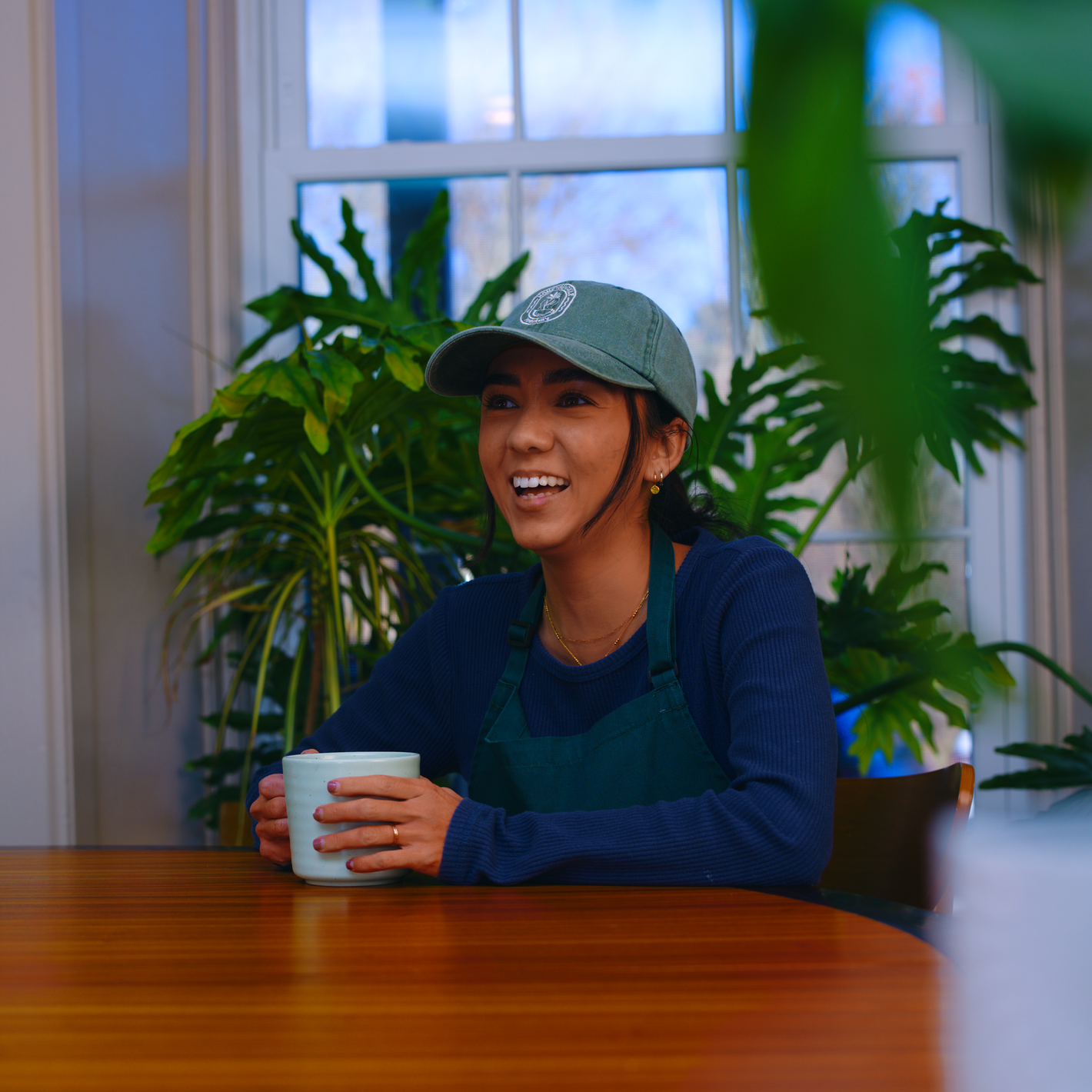 Person smiling at a table, holding a mug in a plant-filled room.