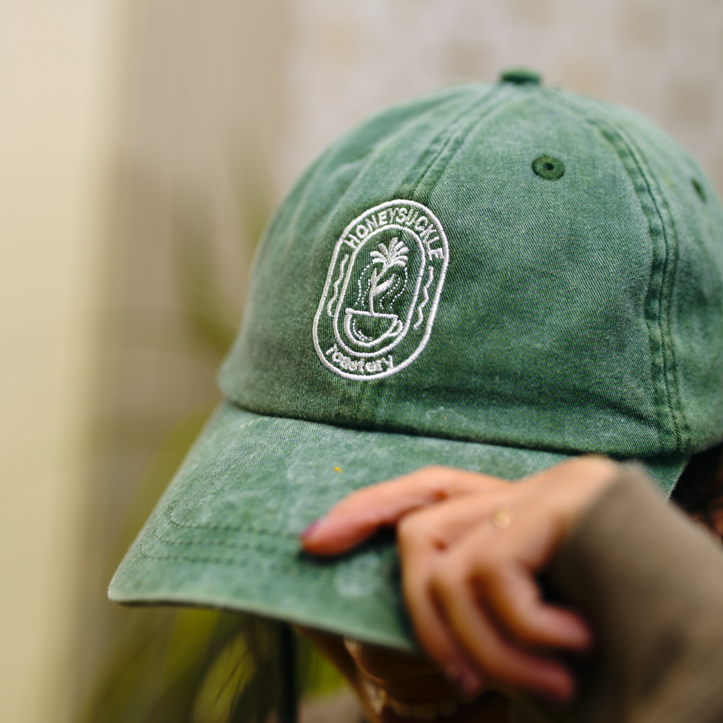 Green baseball cap with white embroidered logo, worn and adjusted by a hand indoors