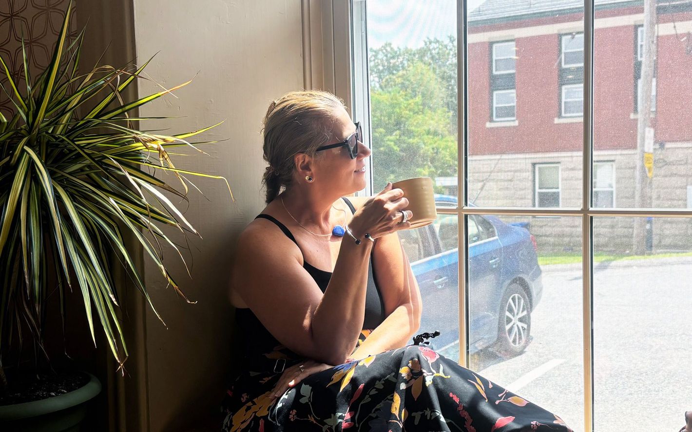 Person seated by a window, holding a mug and looking outside, with a potted plant and parked car nearby.