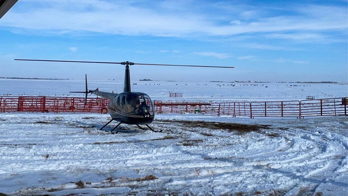 Helicopter parked on a snowy airfield under a clear blue sky, with red fencing in the background
