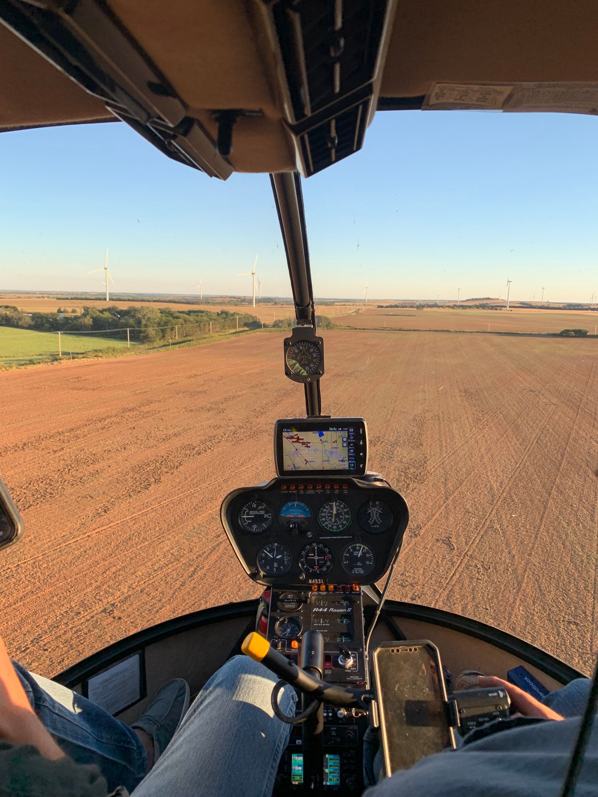 Helicopter cockpit view over a plowed field at sunset, with farmland and horizon ahead