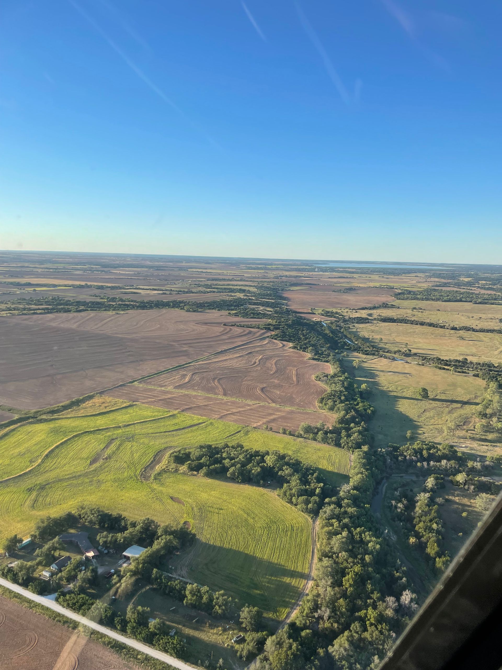 Aerial view of farmland and green fields under a clear blue sky, with a road or tree line dividing the landscape.