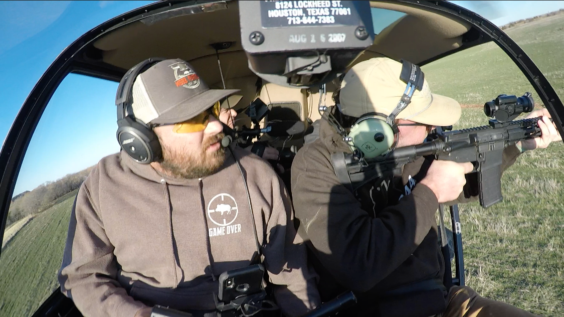 Two men in a helicopter cockpit, one aiming a rifle while wearing headsets and sunglasses.