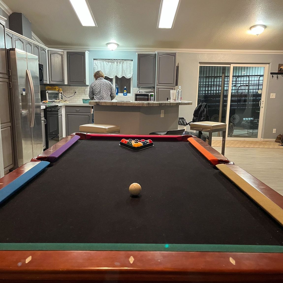Pool table in a bright kitchen; a person stands at the counter in the background.