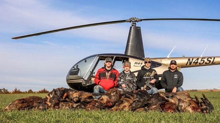 Four people pose beside a helicopter behind a pile of hunted animals in a grassy field.