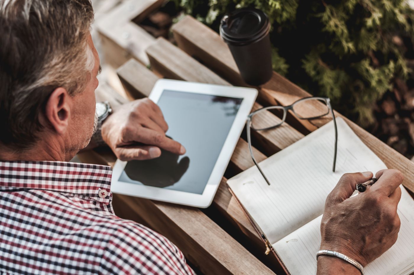 A man is sitting on a bench using a tablet and writing in a notebook.