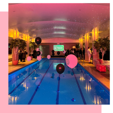 Indoor pool with black and pink balloons, illuminated ceiling, and people gathered.