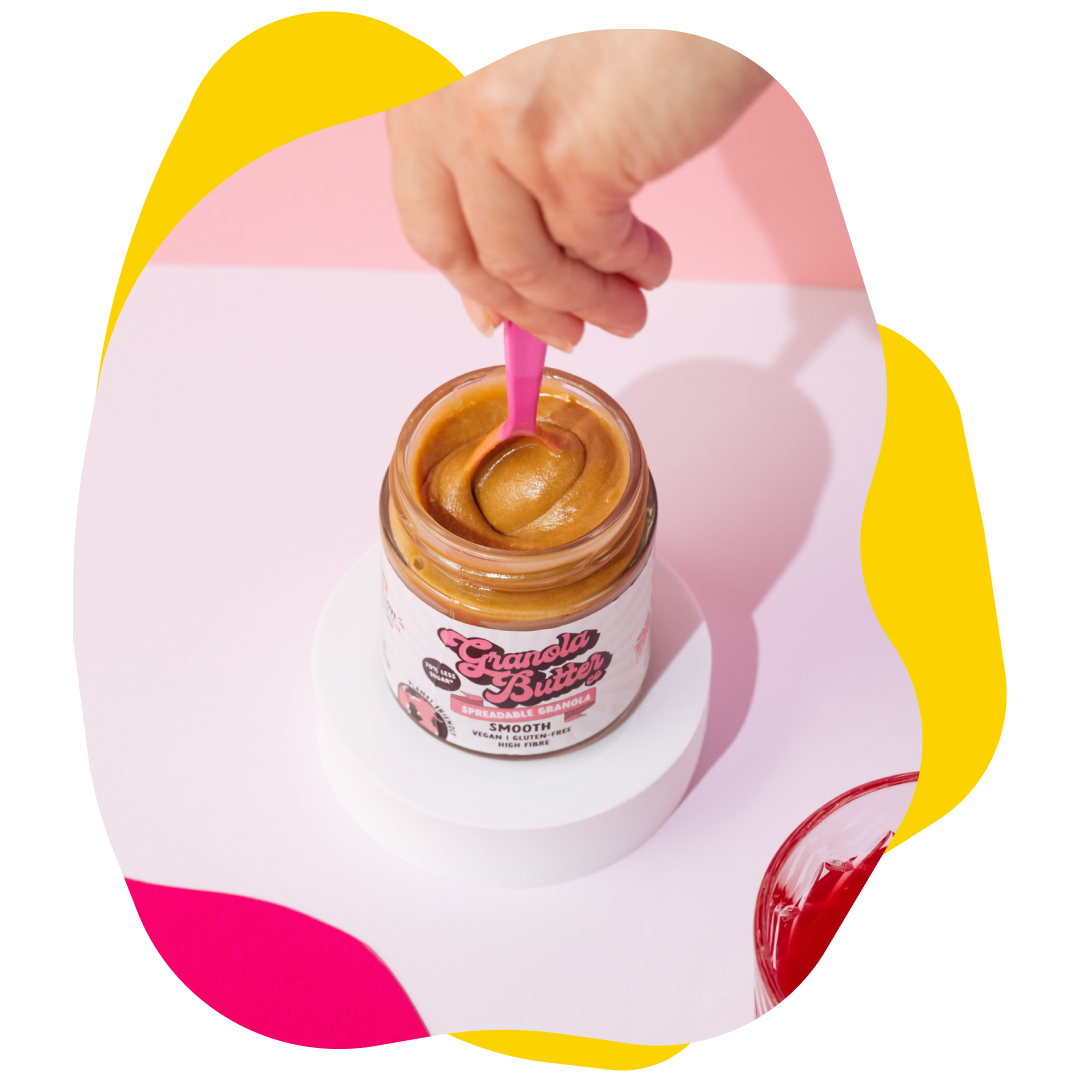 Hand scooping nut butter from a jar with a pink spoon, set on a white stand. Bright background.