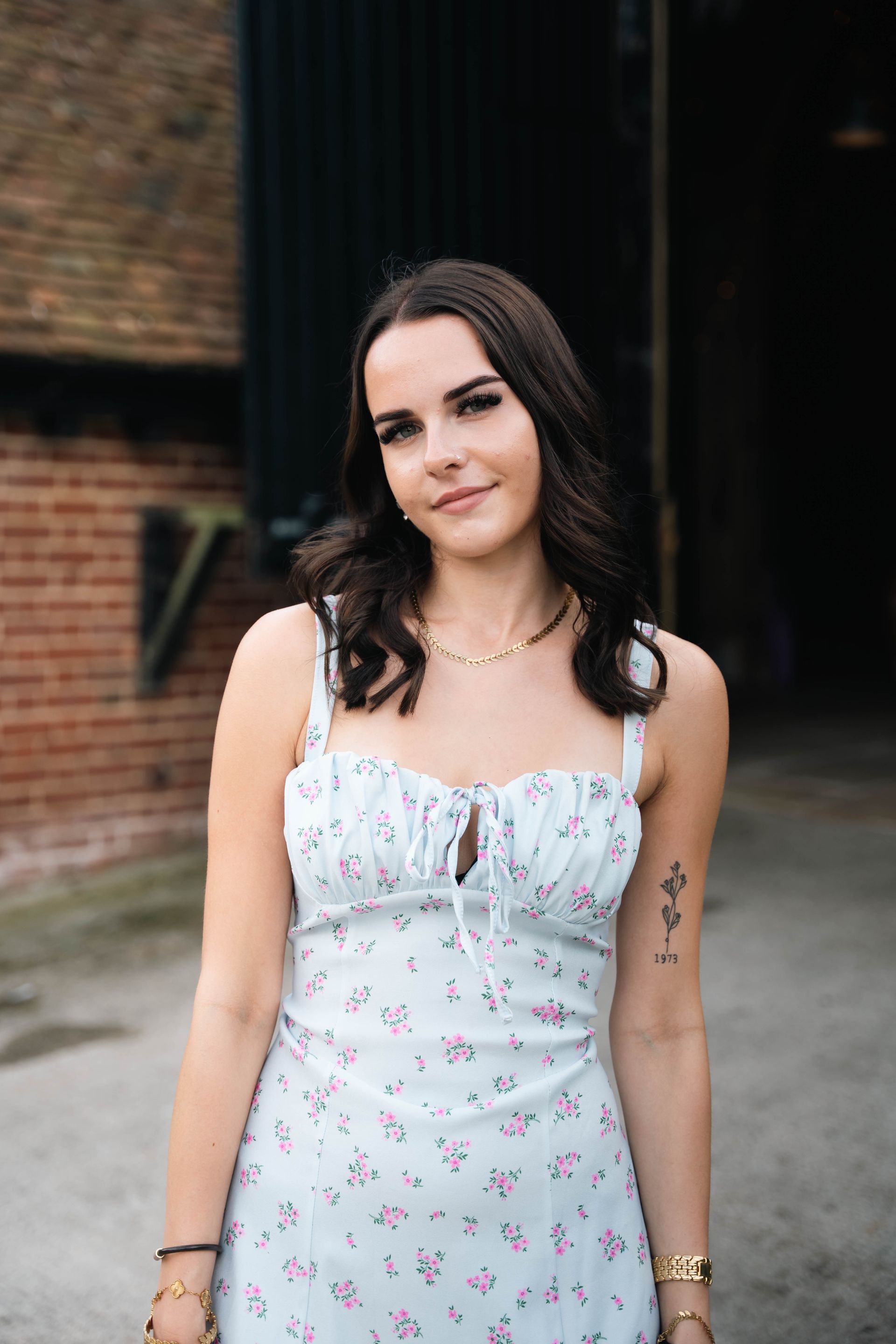 Woman in a light blue floral dress, smiling, standing in front of a brick building entrance.