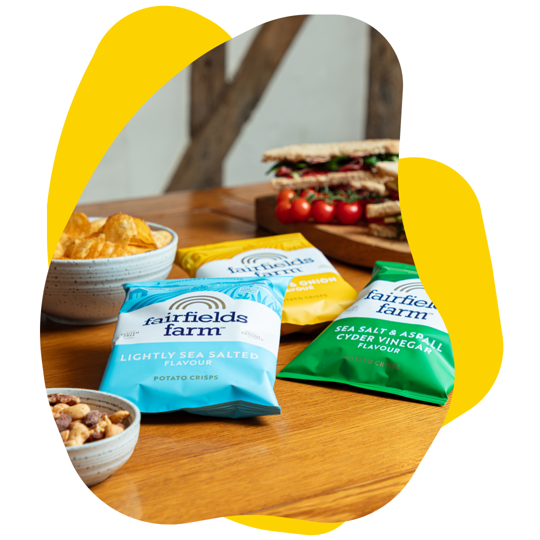 Bags of Fairfield Farm crisps and a bowl of crisps and nuts on a table with a sandwich.