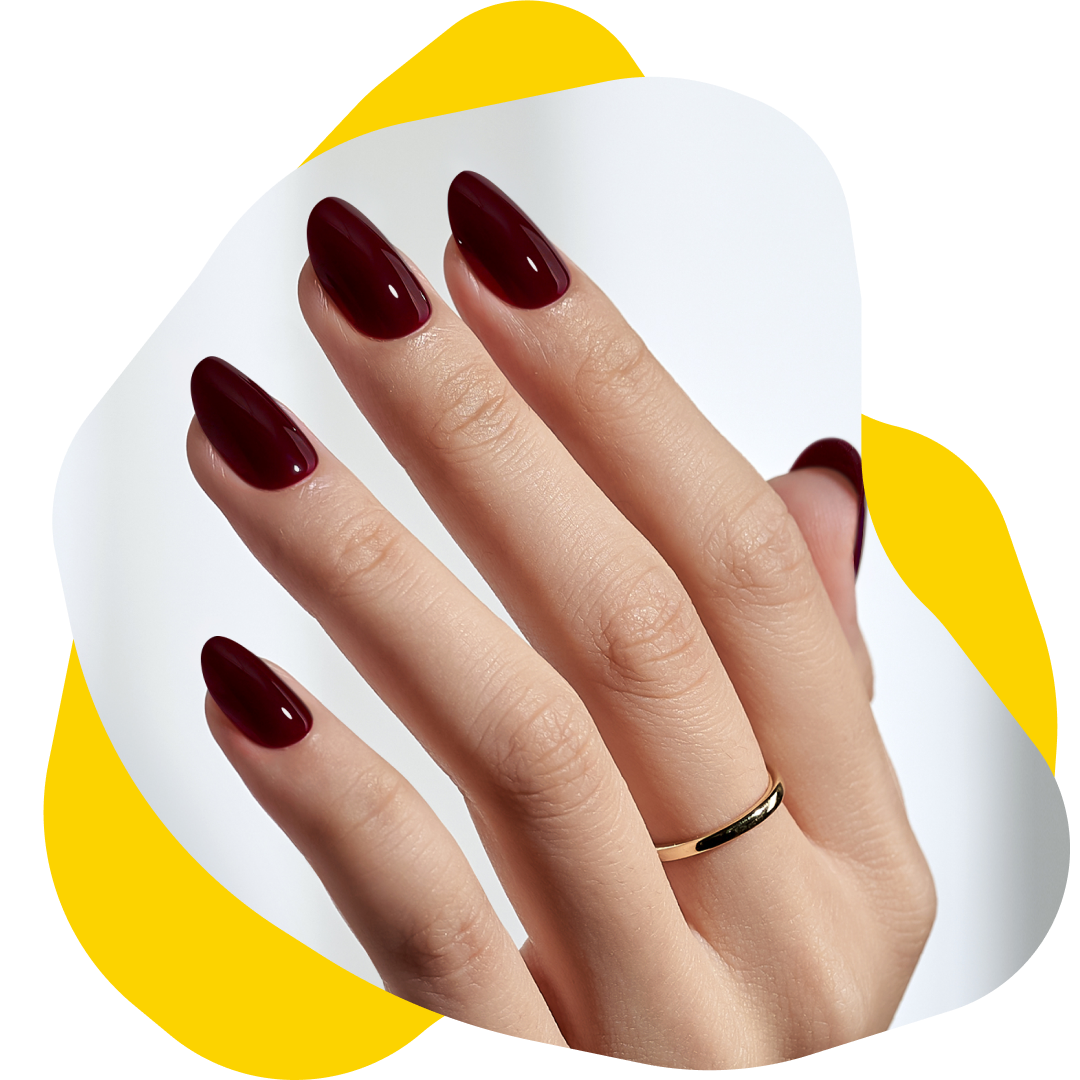 Hand with polished, maroon-colored almond nails; gold ring; resting on a white and yellow background.