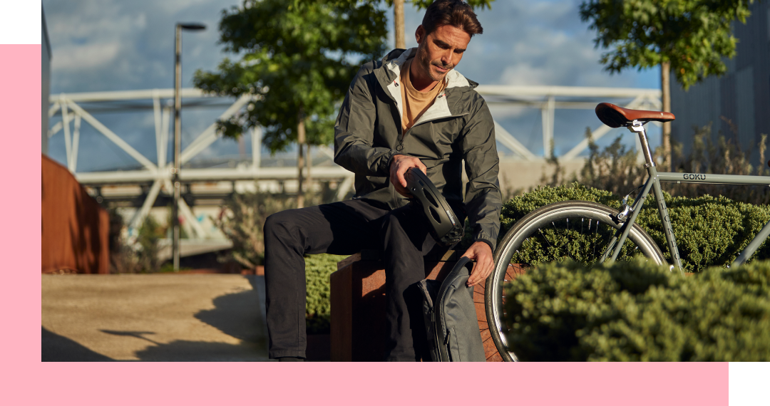 Man sitting on bench adjusts bag near bicycle. Outdoors, neutral-toned clothing, trees, modern bridge.