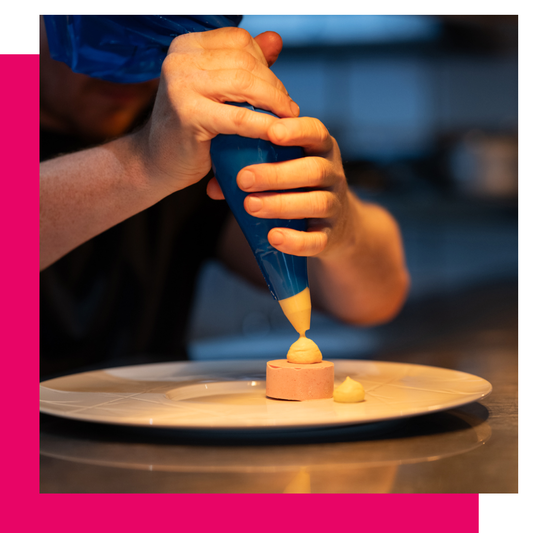 Chef decorates food on a plate with cream from a piping bag in a kitchen.