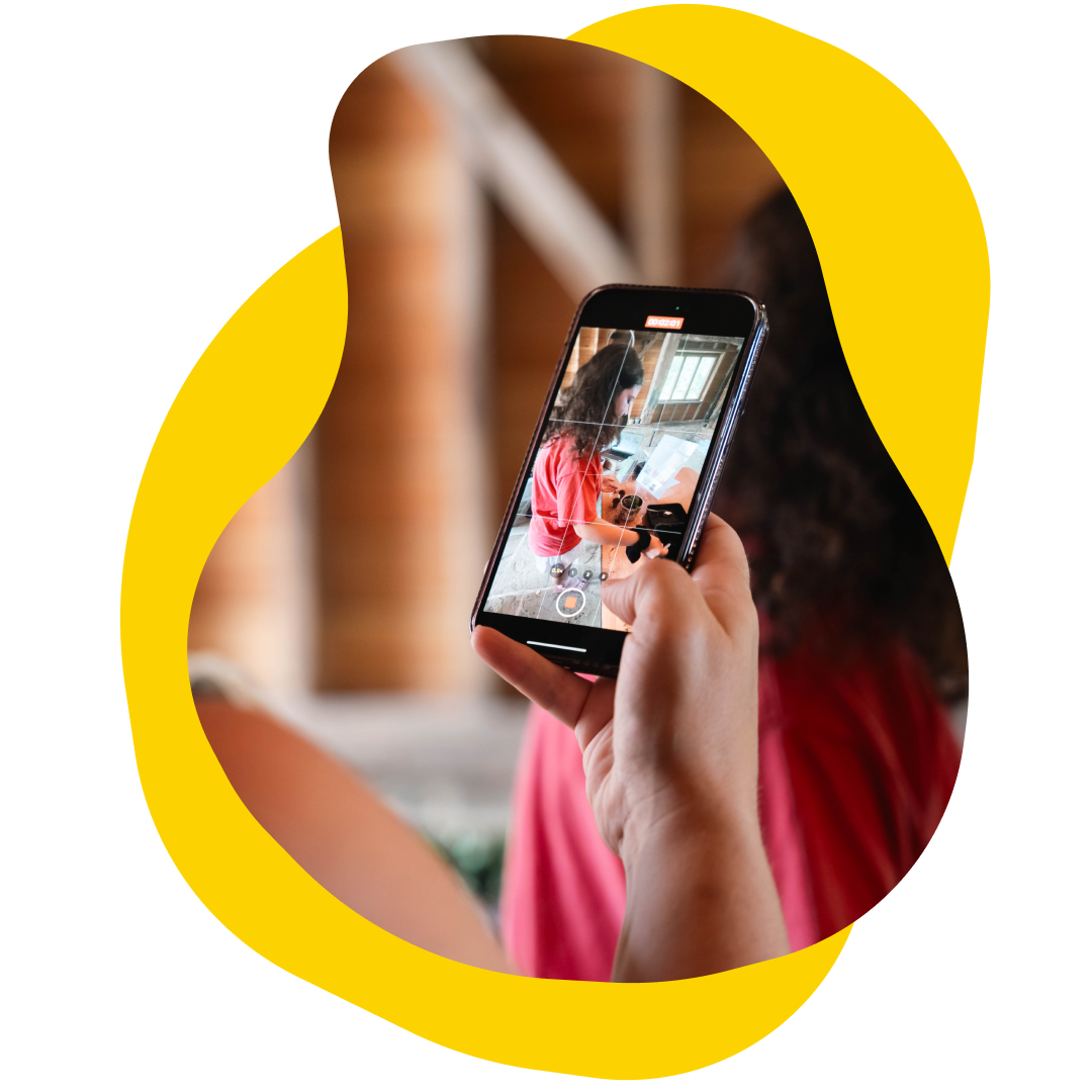 Person holding a smartphone, recording video of someone indoors, with a yellow abstract shape around the image.
