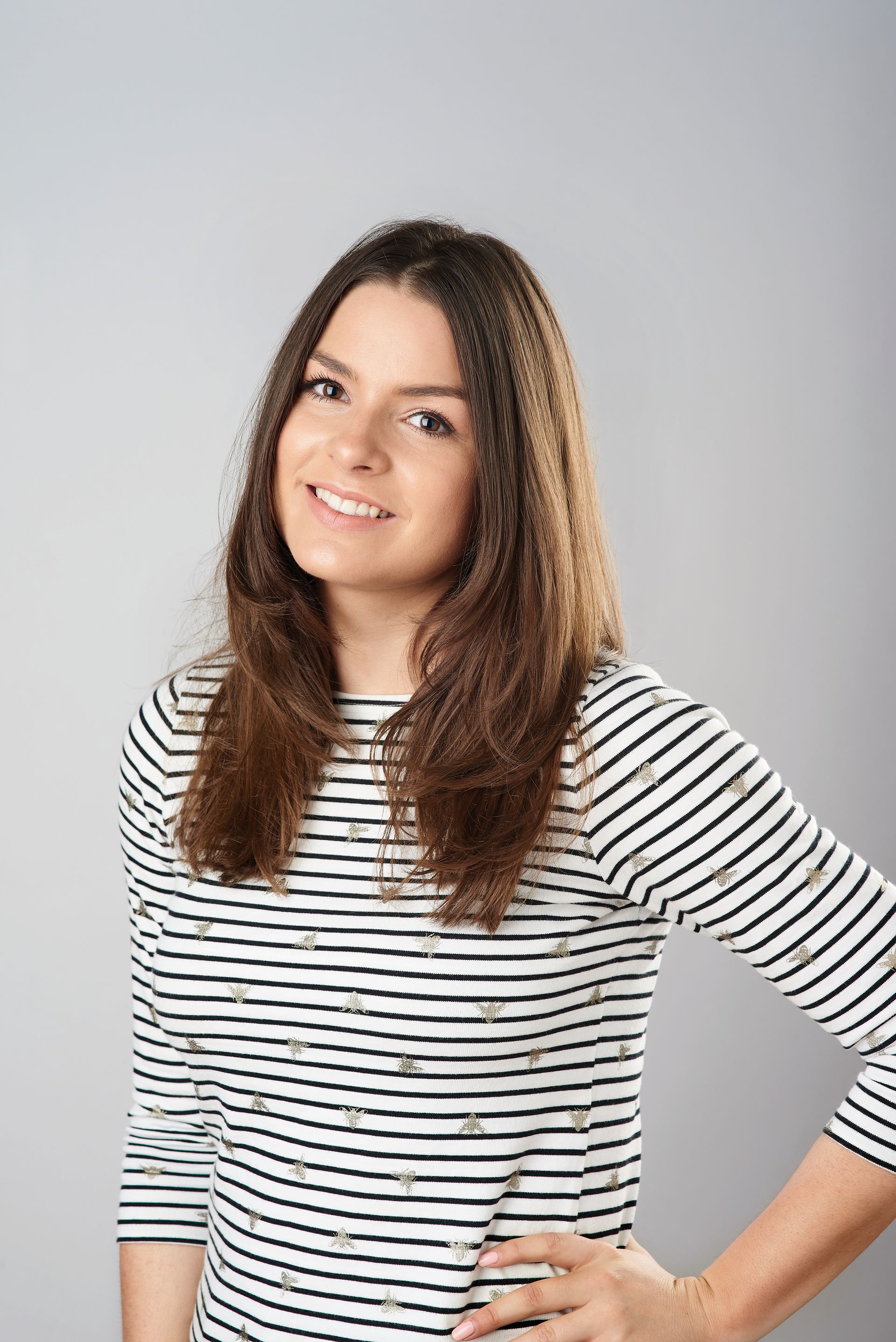 Woman smiling, wearing a striped shirt, hand on hip, against a gray background.