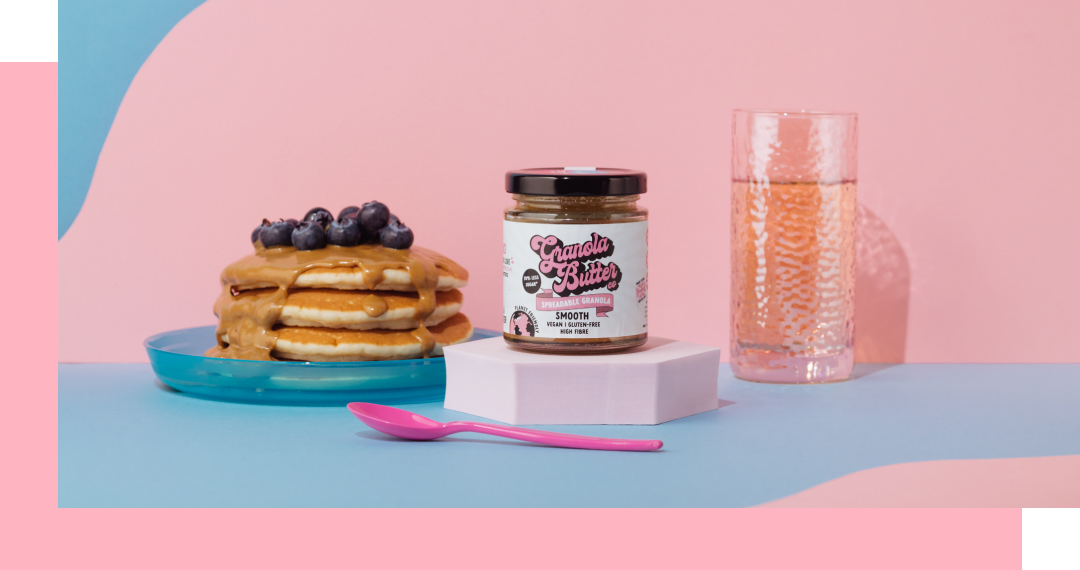 Stack of pancakes with blueberries and peanut butter, jar of peanut butter, a glass of pink liquid, and a pink spoon.