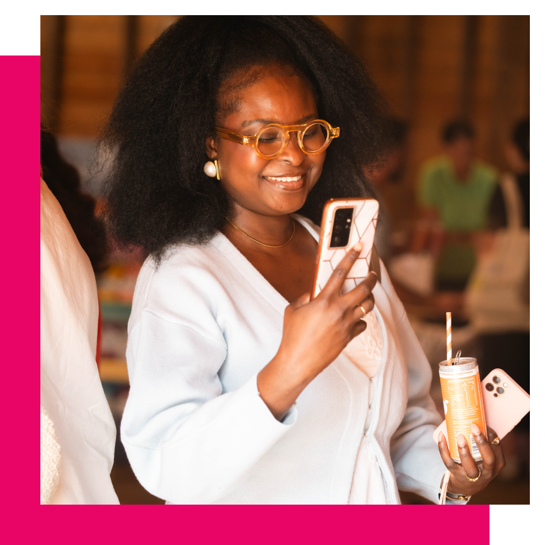 Woman in glasses smiles while holding a phone and drink, taking a photo.