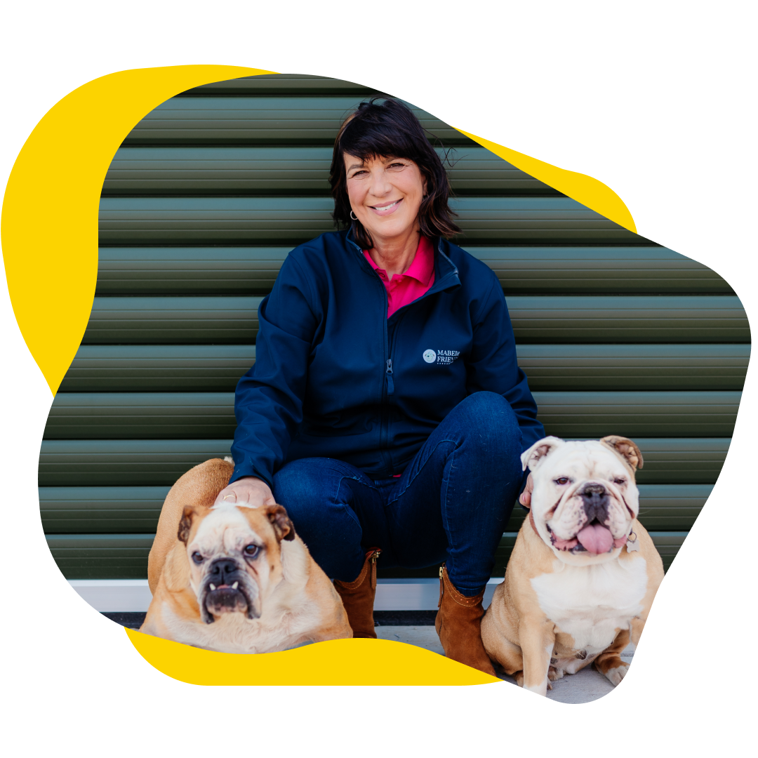 Woman crouches with two bulldogs; one petted, all smile. Blue jacket, jeans, brown boots, setting is green ribbed.
