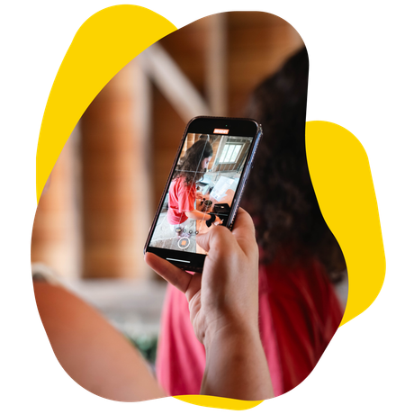 Person holding a phone, filming another person. The setting is indoors, with a bright yellow background.