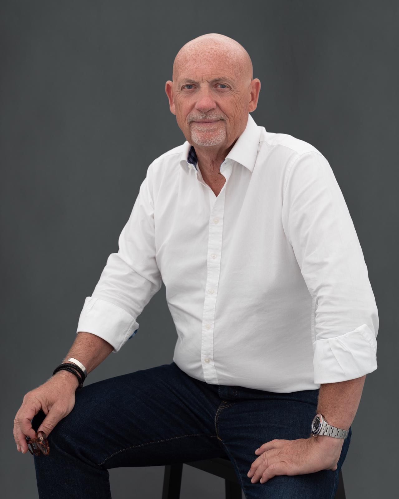 Man in white shirt and jeans sits on a stool, looking at the camera. Gray background.