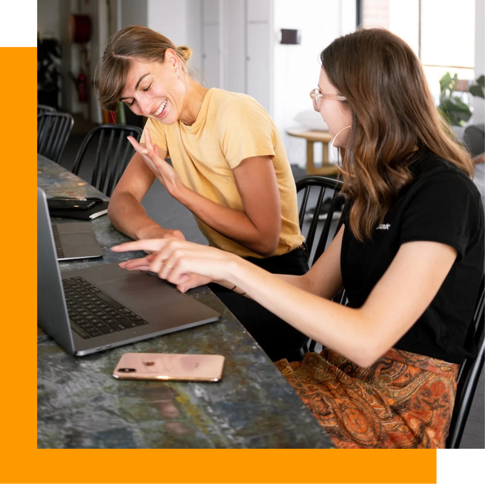 Two women smiling, looking at laptop screen at a table. One points while the other gestures.