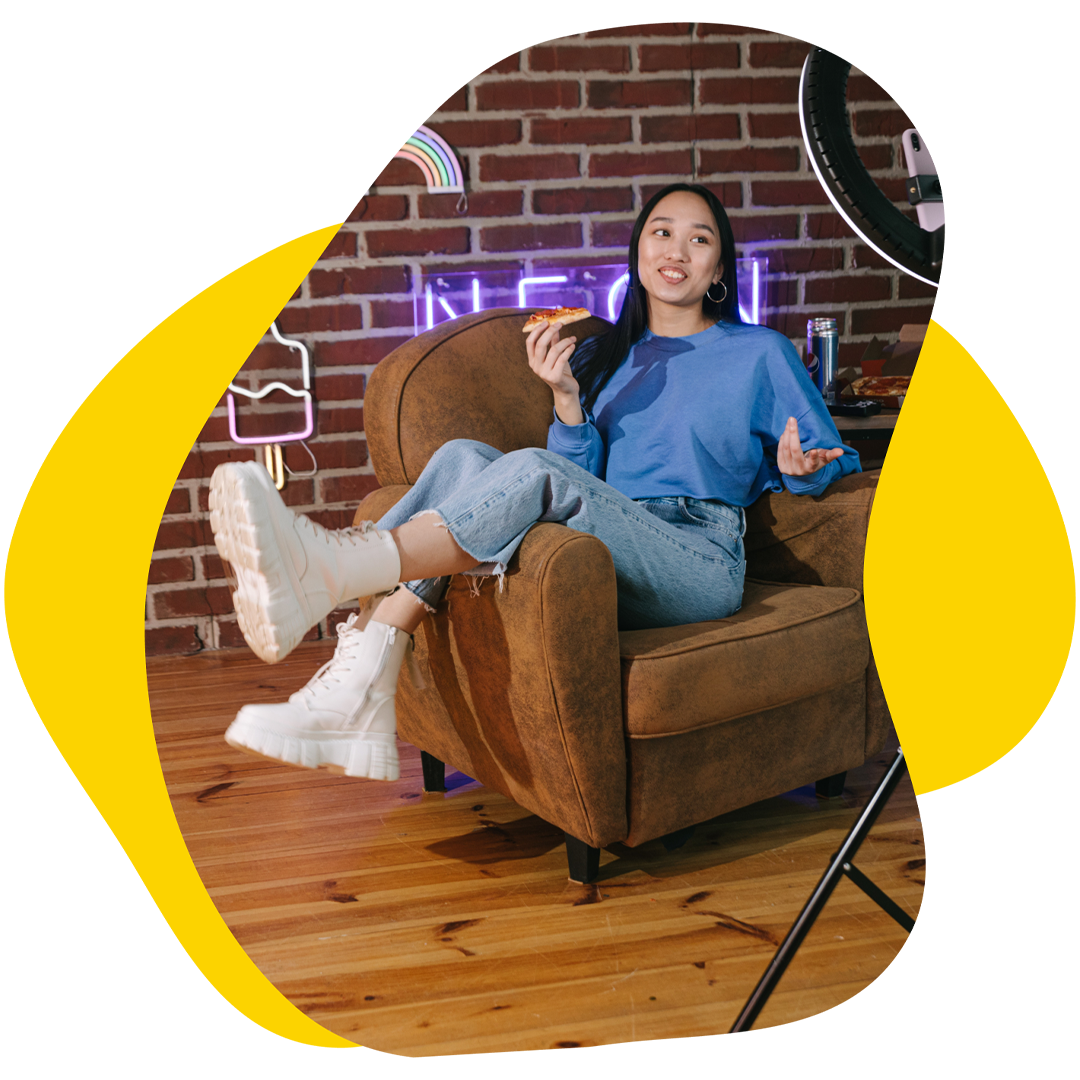 Woman sitting in armchair eats pizza, filming a video. Brick wall with neon signs.