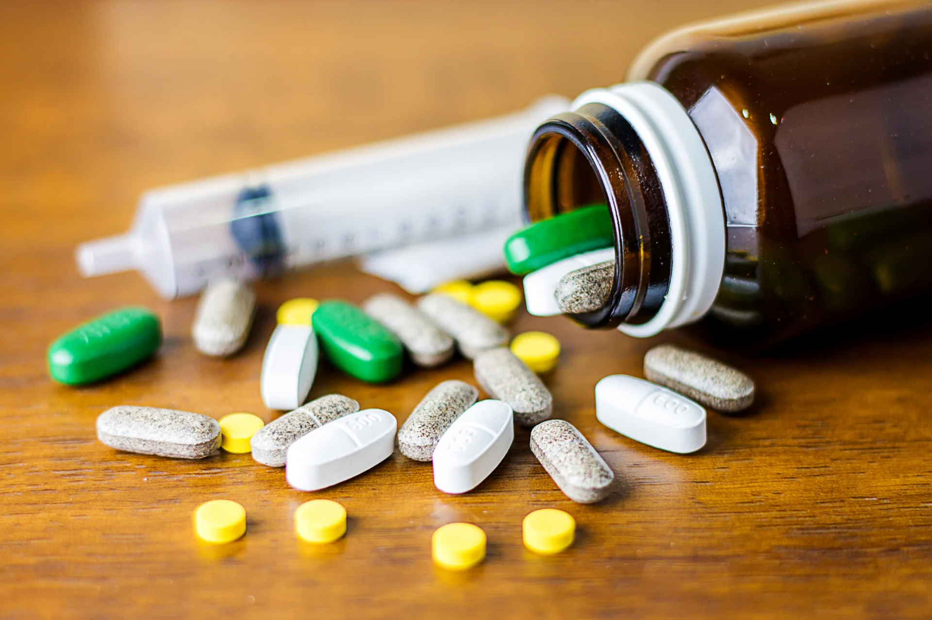 Pills of various colors spilling from a bottle next to a syringe on a wooden surface.