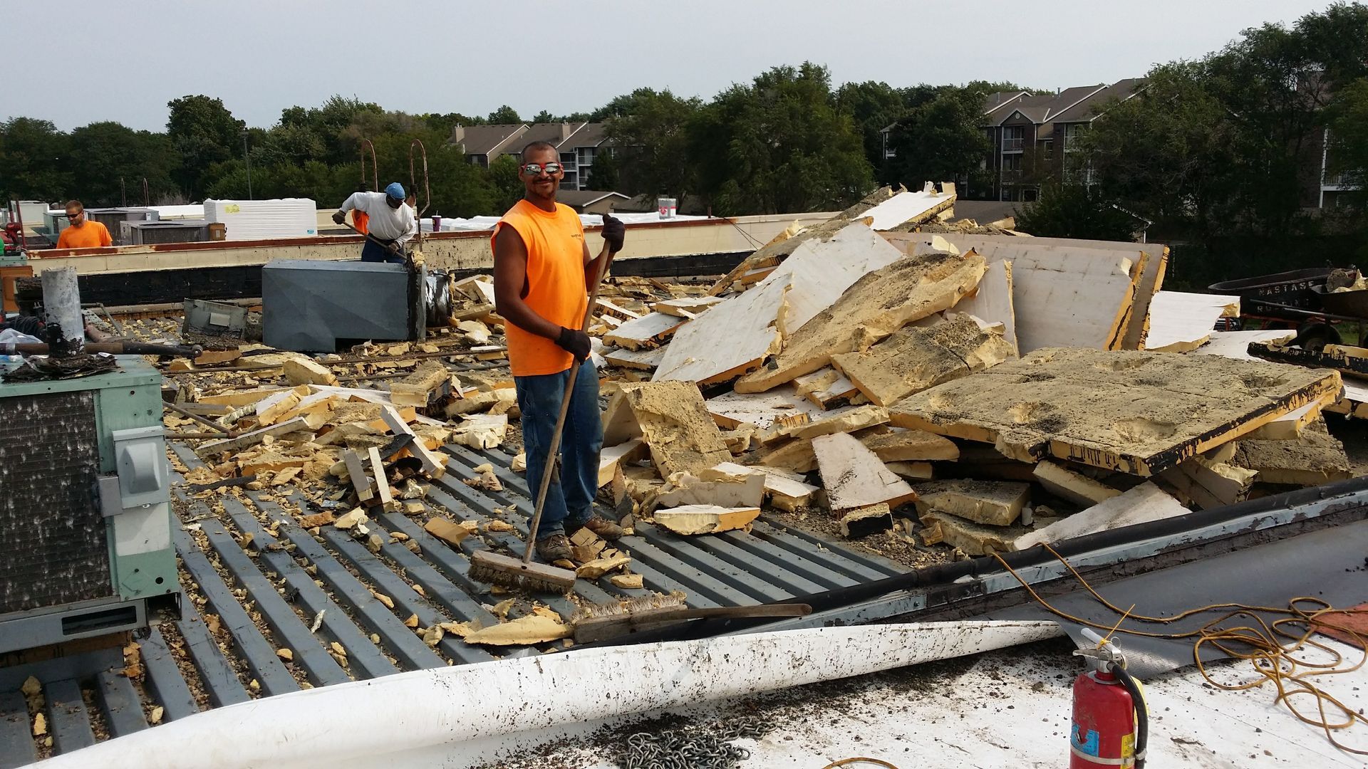 Construction workers in bright safety vests work on a flat roof strewn with large piles of insulation debris.