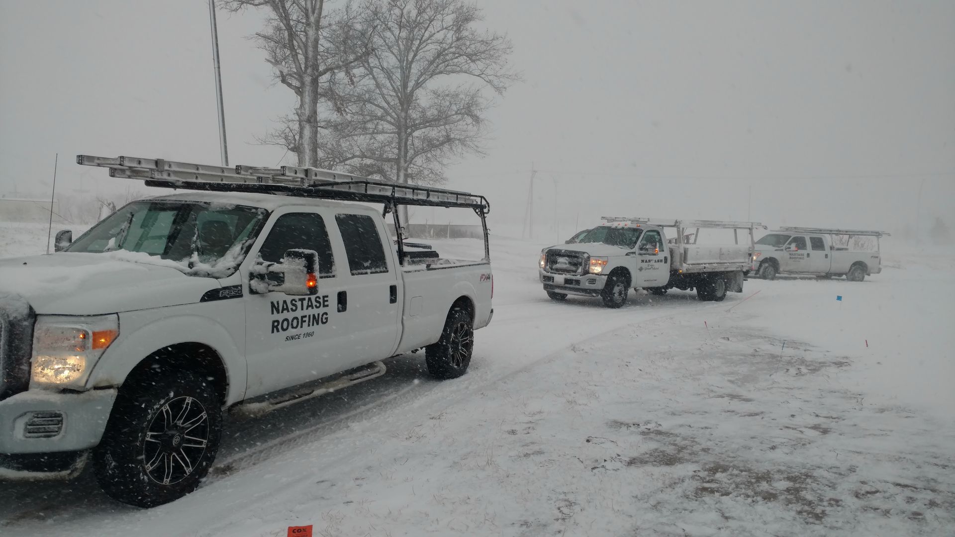 Three white utility trucks with ladder racks are parked on a snow-covered field during a heavy snowstorm.