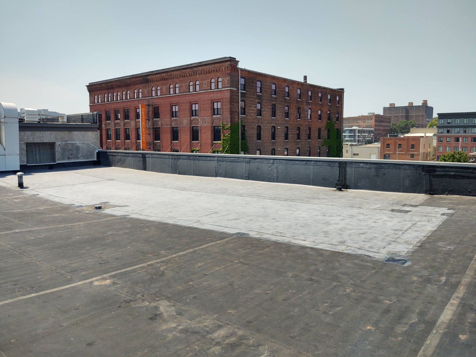 A view from a flat, black rooftop looking across at a multi-story red brick industrial building under a clear blue sky.