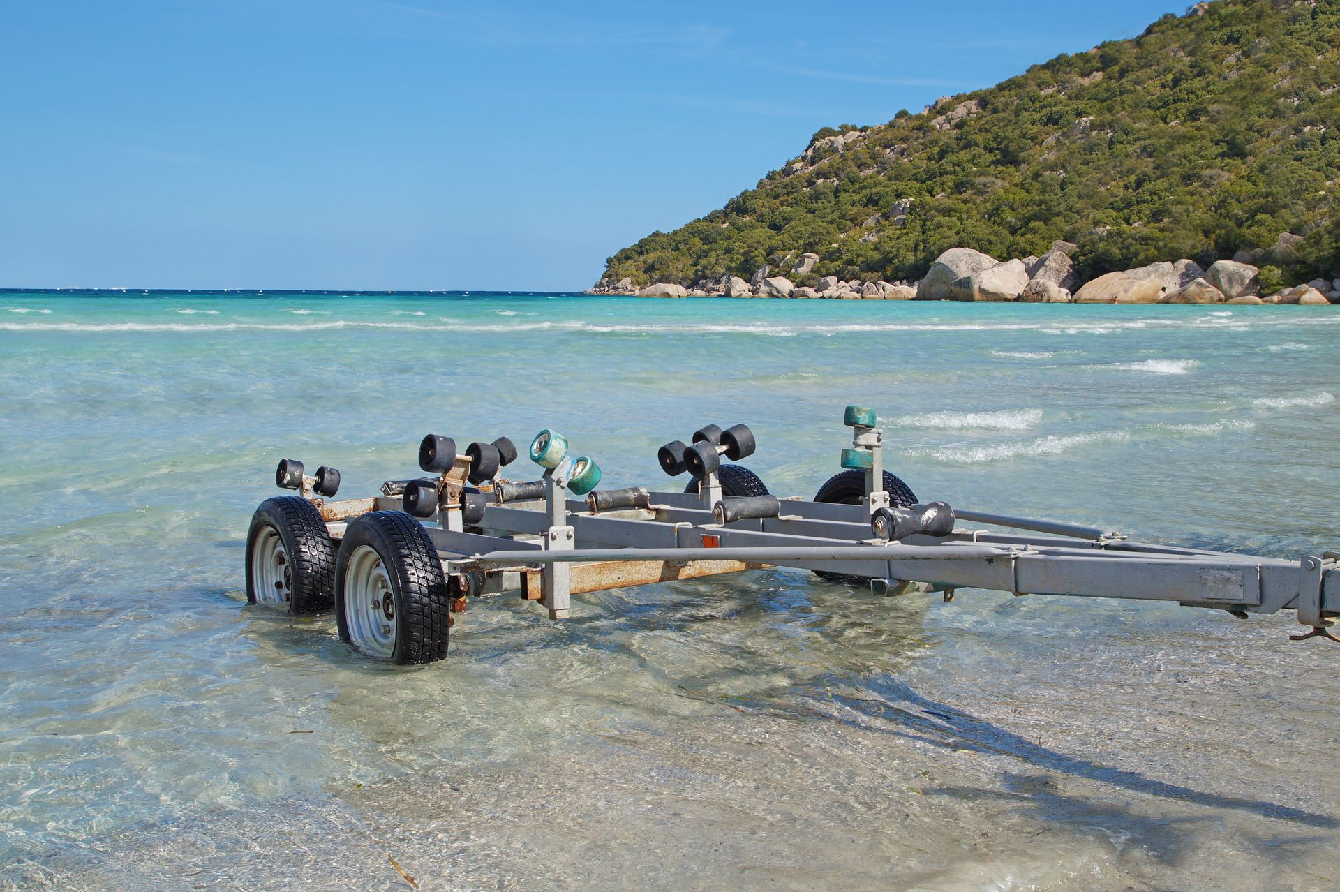 Boat trailer on a shallow turquoise beach with a rocky, green hill in the background