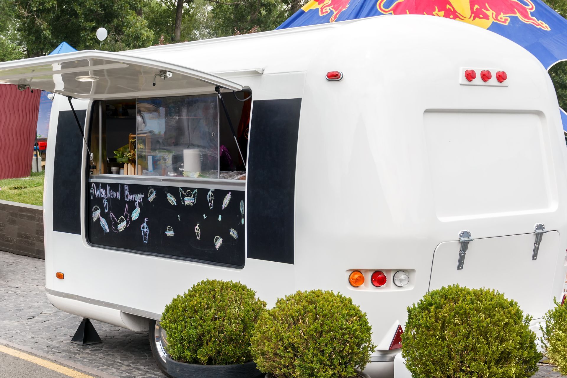 White food trailer with open serving window and black awning, parked outdoors with shrubs in front