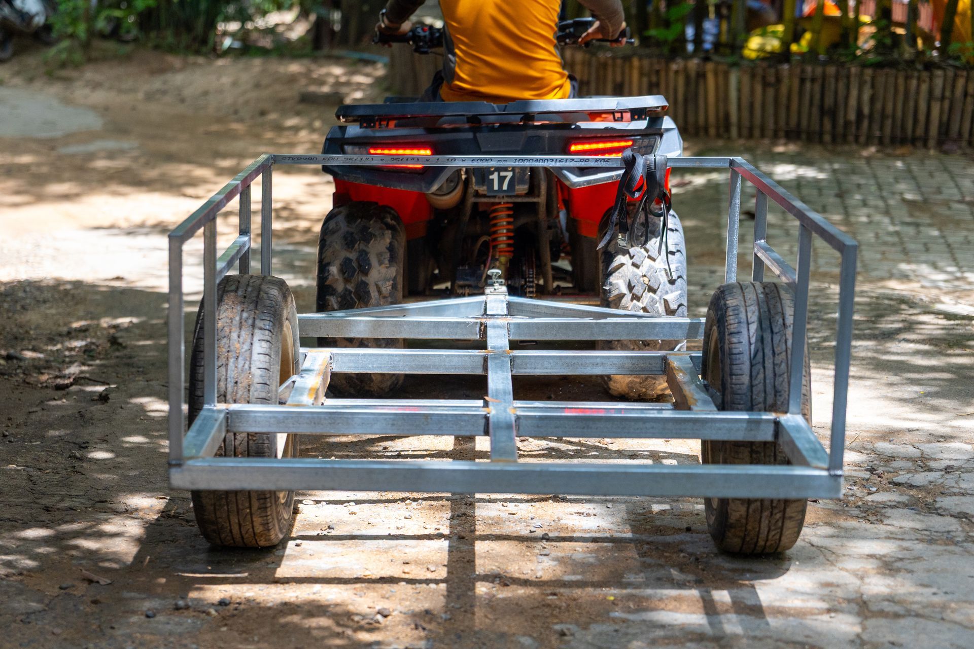 ATV towing a gray metal trailer on a dirt path, with a person in an orange shirt riding ahead