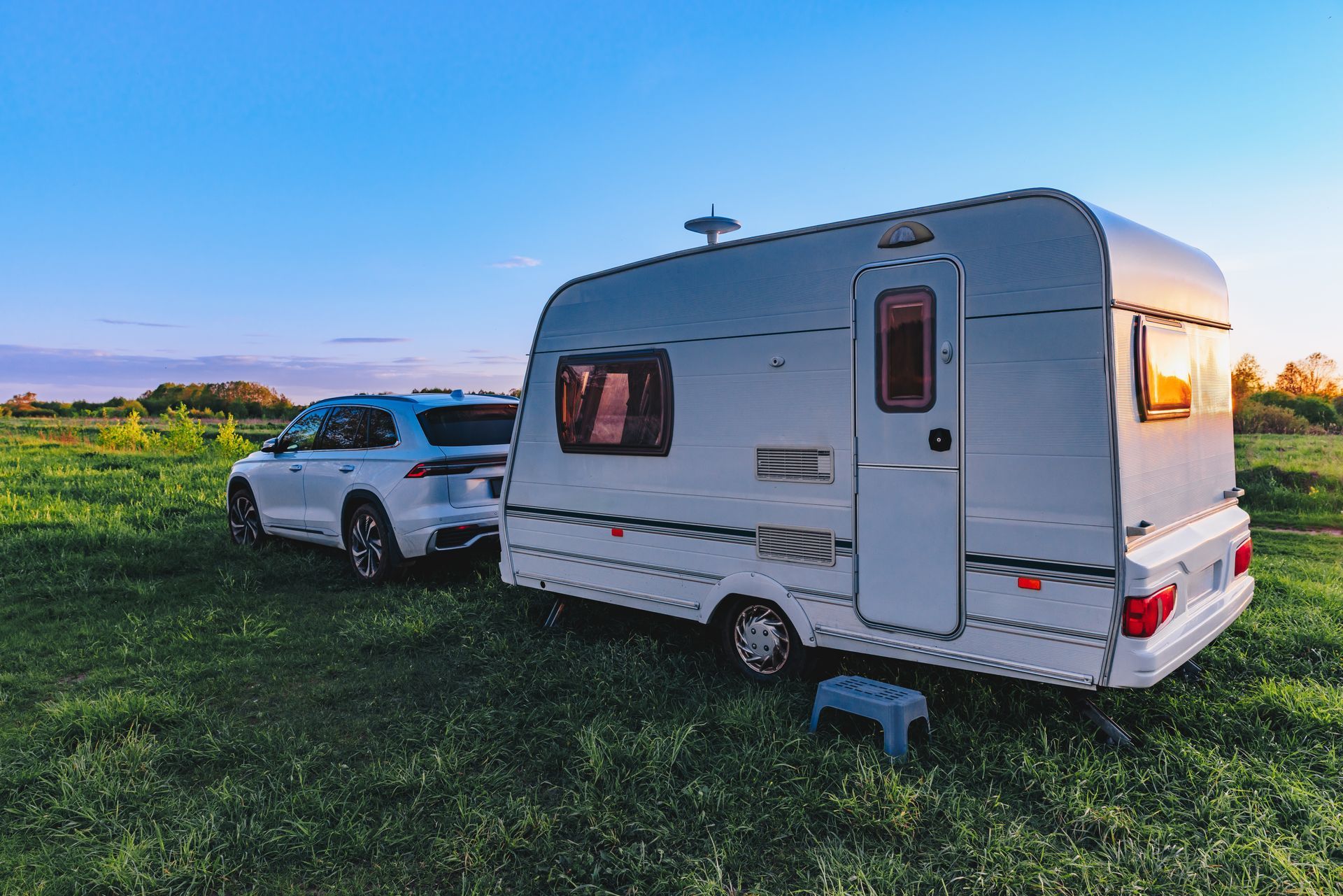 White SUV parked beside a white travel trailer in a grassy field at sunset.