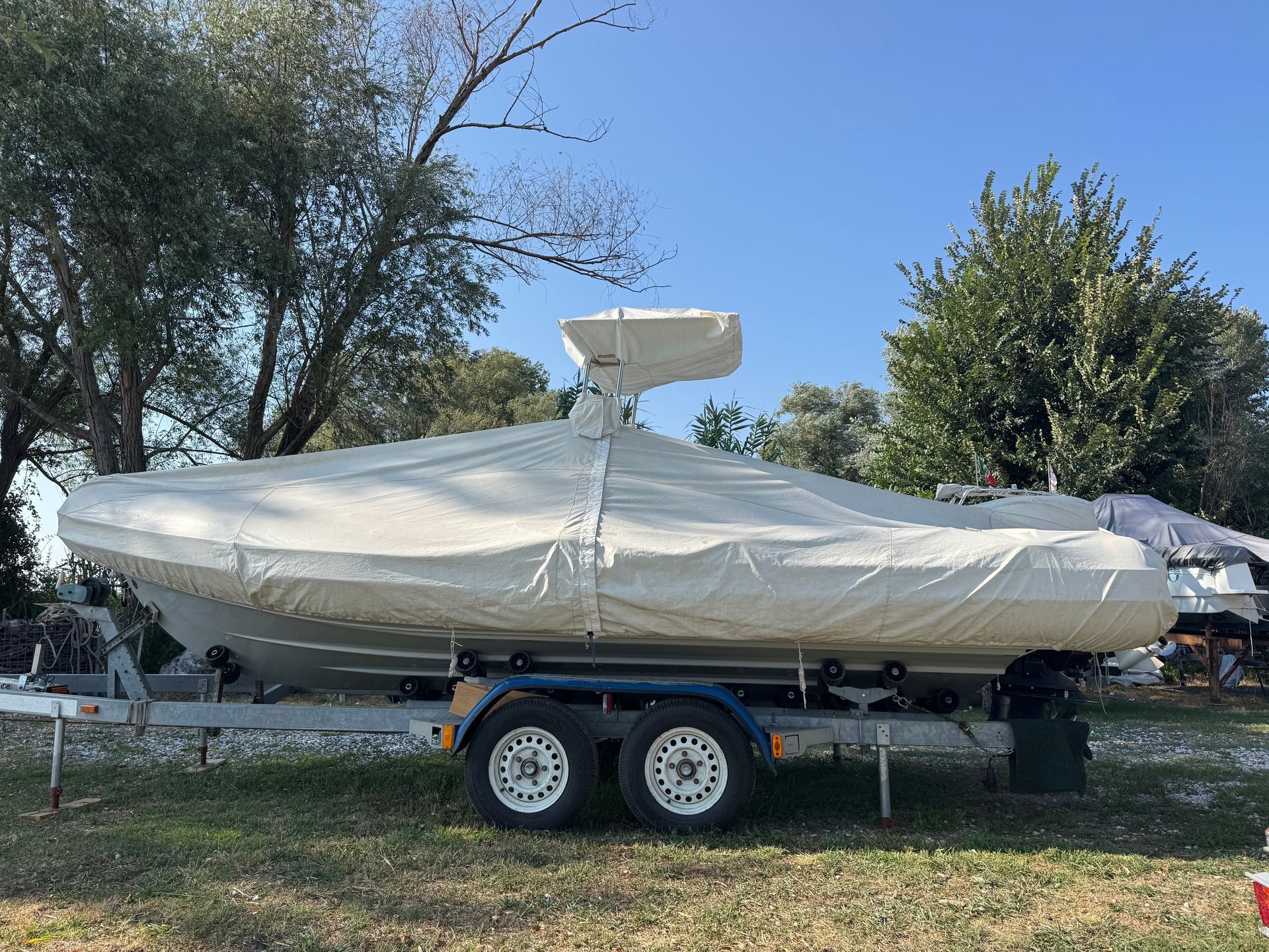 Covered white boat on a trailer outdoors beside trees under a clear blue sky