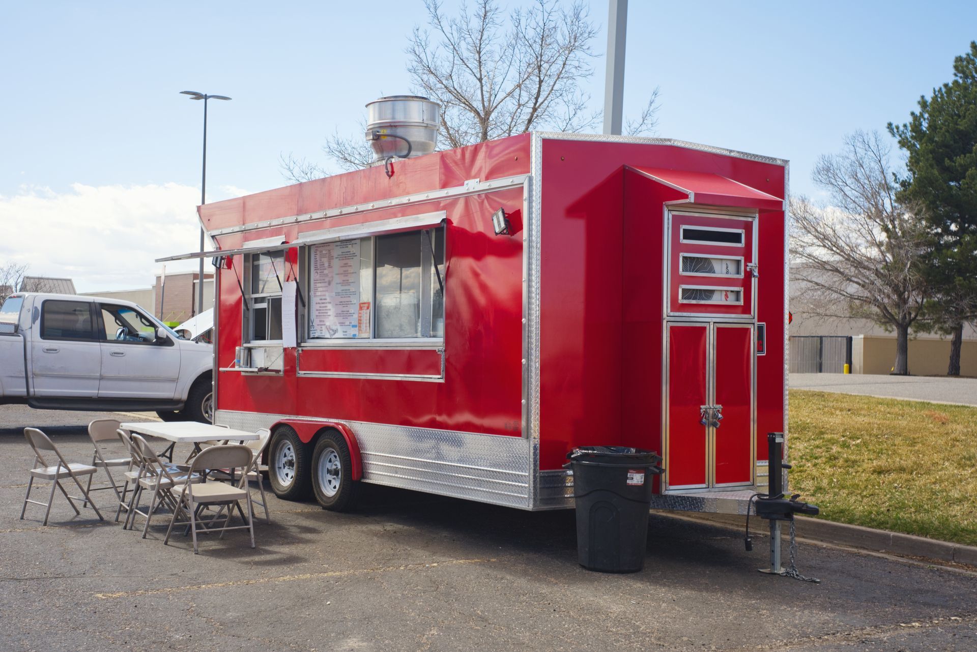 Red food truck with serving window, picnic tables, and a white pickup parked beside it outdoors.