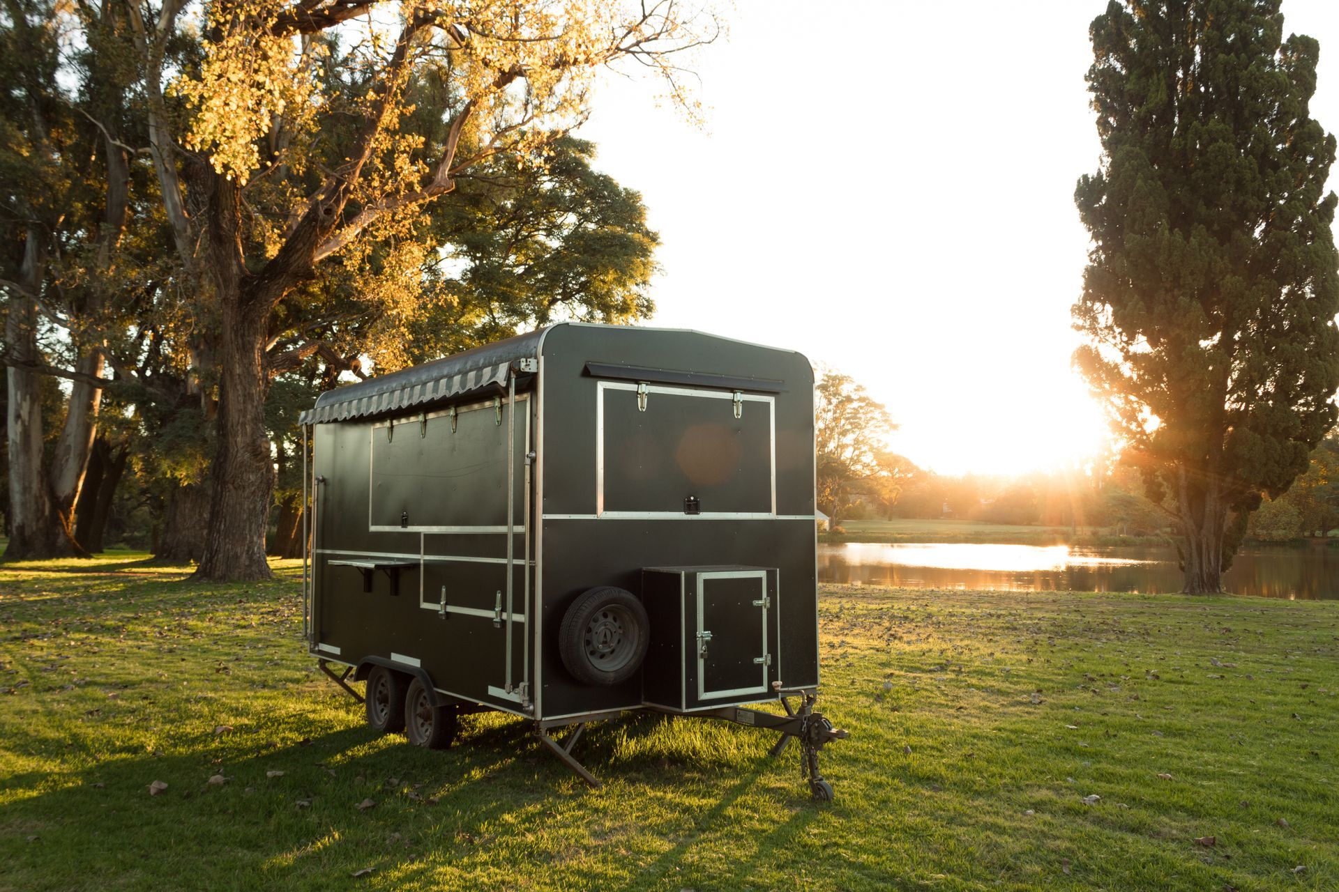 Black camper trailer on grass at sunset beside trees and a lake