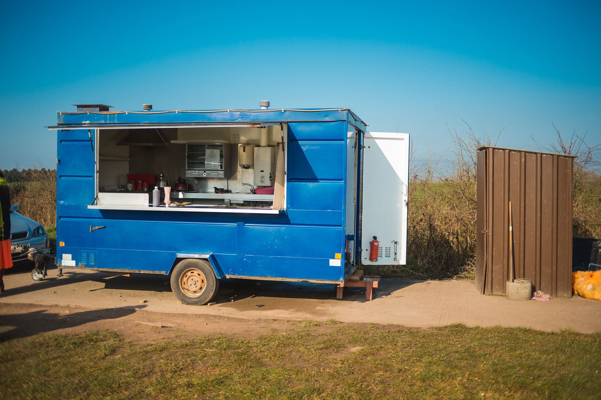 Blue food truck parked on a dirt lot beside a brown fence, with a service window open.