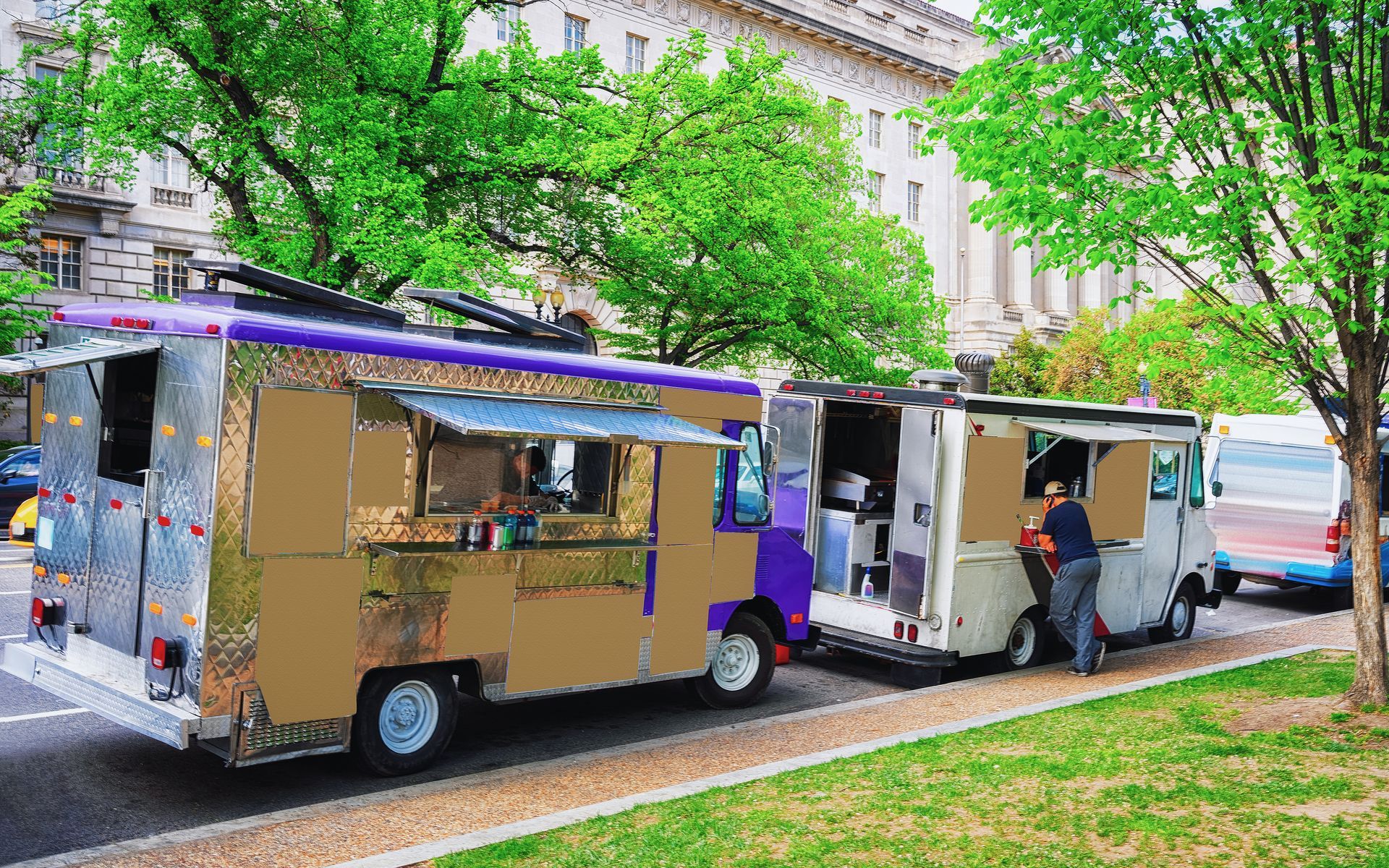 Two food trucks parked along a tree-lined street, one serving a person at the side window.