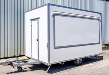 Small white trailer kiosk with large serving window, parked on pavement beside a corrugated metal wall.