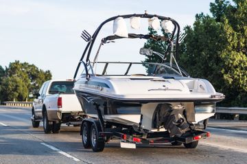 White speedboat on a trailer behind a white SUV on a road, with trees in the background