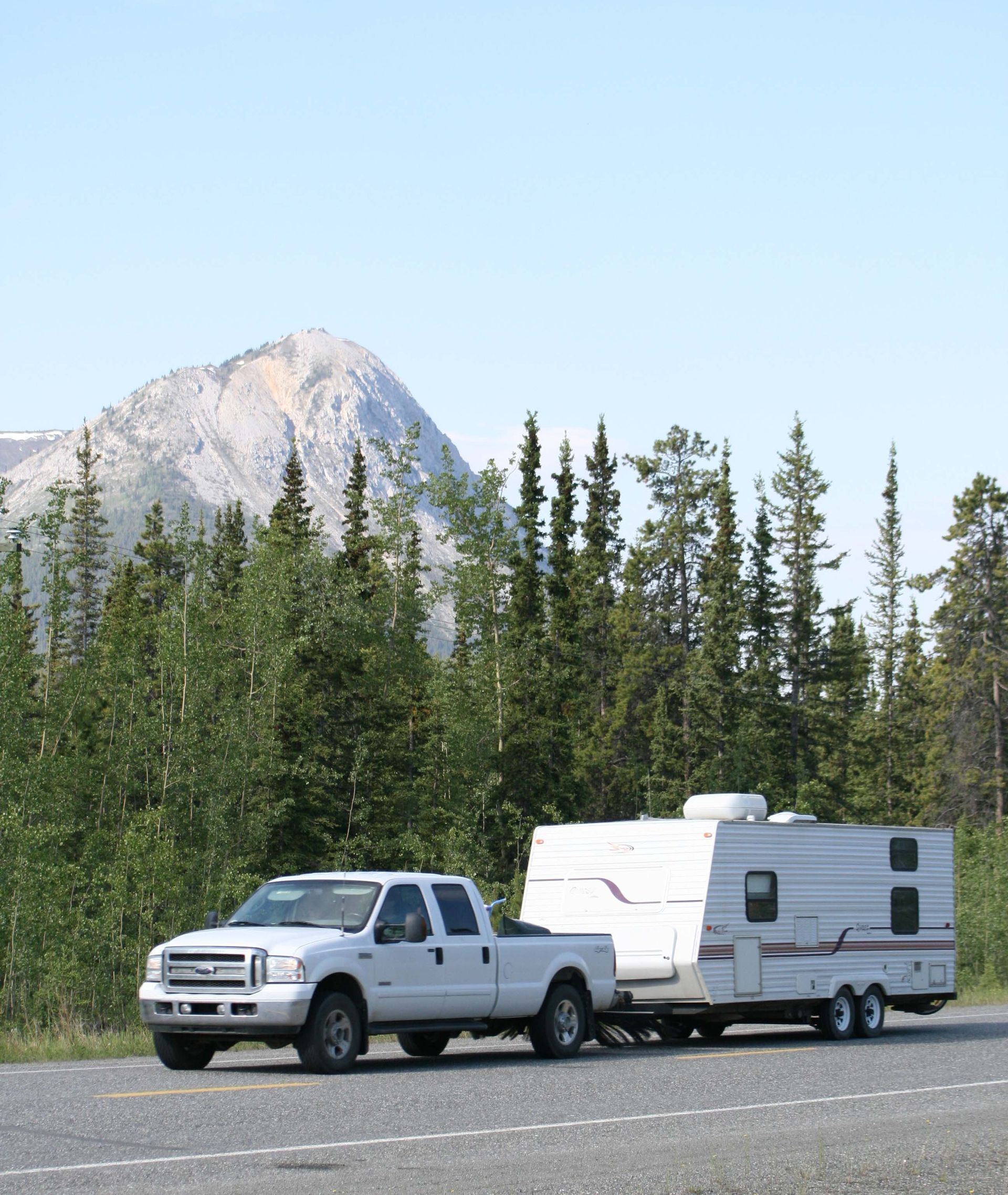 White pickup truck towing a white camper trailer on a road with pine trees and a mountain in the background