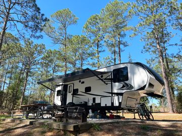 White and black fifth-wheel RV parked at a campsite among tall pine trees under a blue sky.