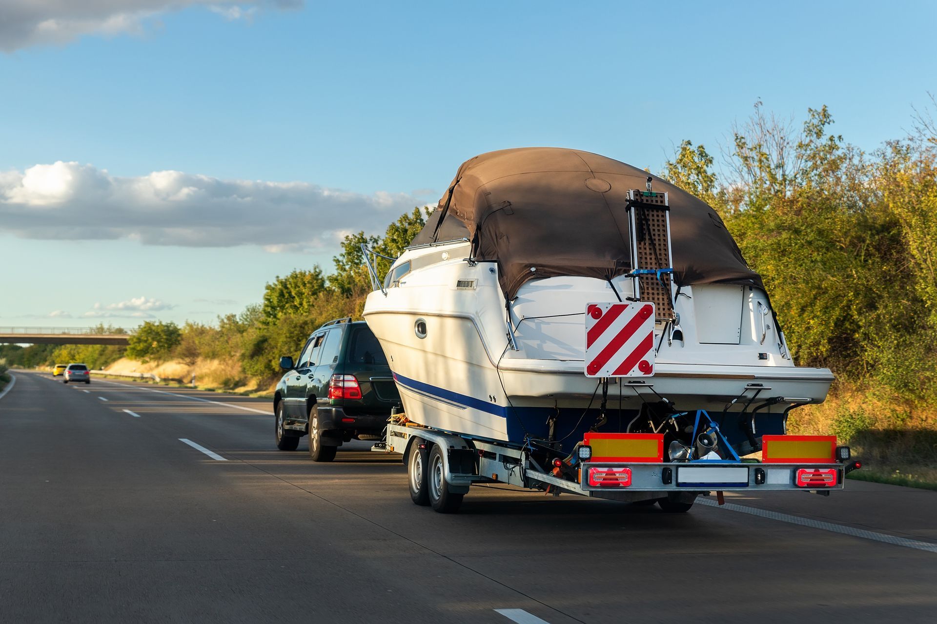 Boat on a trailer being towed on a highway behind a black vehicle under a partly cloudy sky