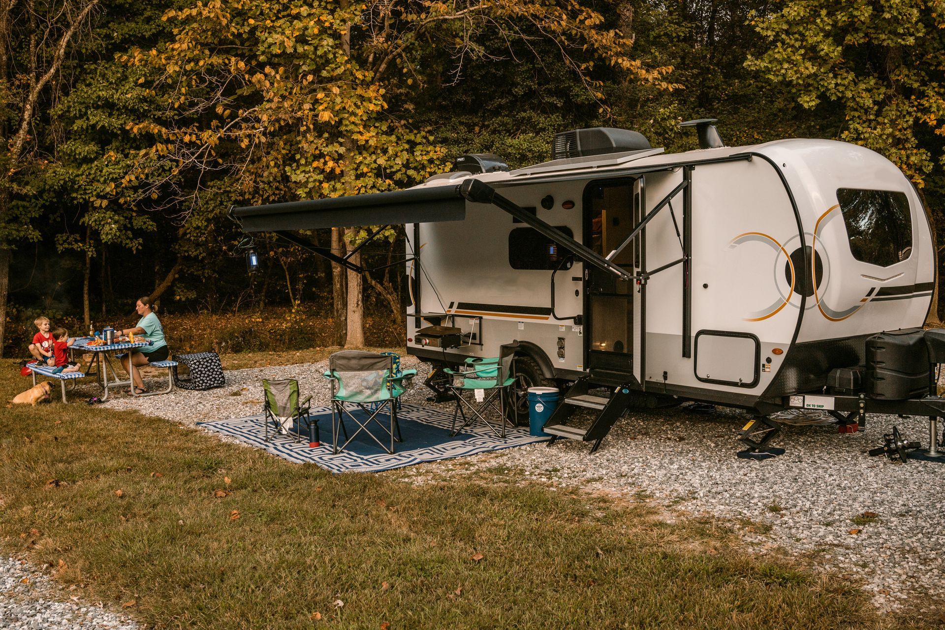 Camper trailer at a wooded campsite with an awning, picnic table, and chairs on gravel and grass