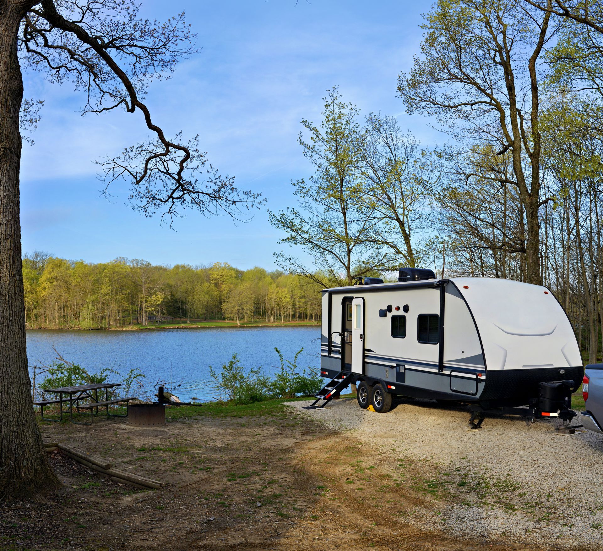 Small camper trailer parked by a lake with trees and a blue sky in the background