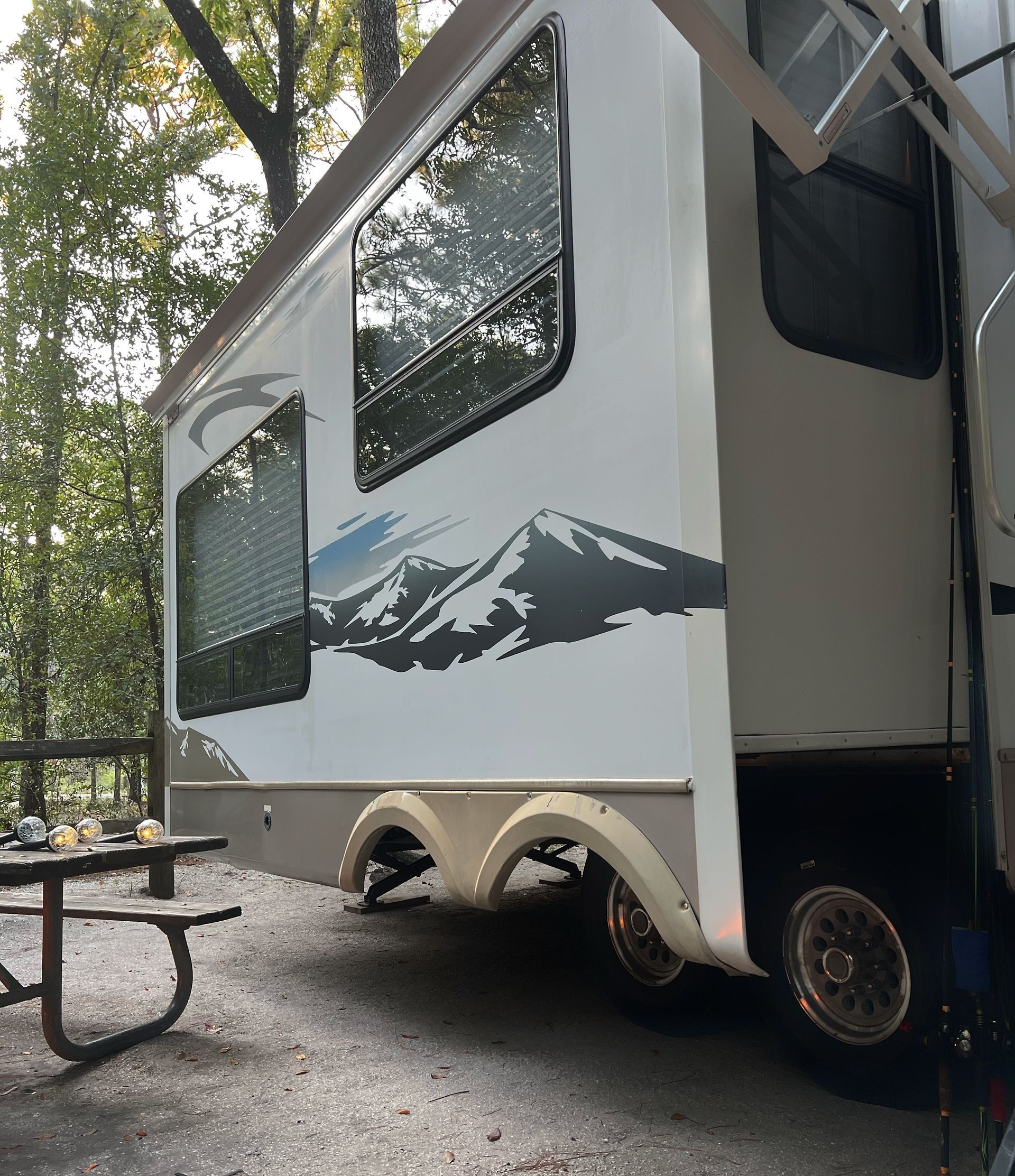 Small white camper trailer with mountain mural parked at a wooded campsite beside a picnic table.