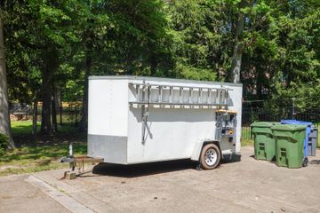 White utility trailer parked outdoors beside green trash bins near trees, in a sunny paved area